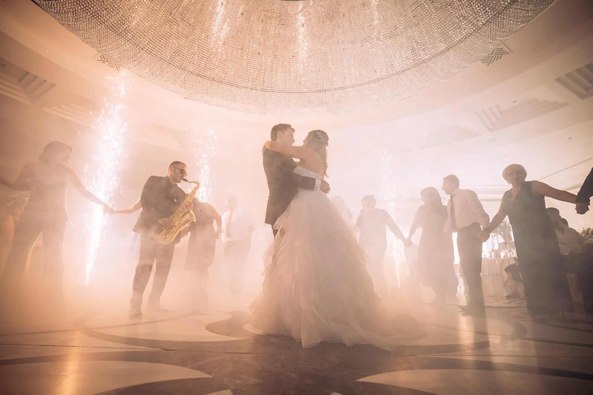 Newlyweds' first dance under a chandelier, surrounded by guests, a saxophonist and cold sparks